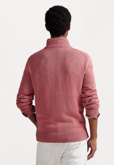 Ροζ πλεκτή ζακέτα με ψηλό λαιμό, ριχτές μανσέτες και στρίφωμα. Το ύφασμα έχει μια διακριτική υφή και φοριέται πάνω από ένα ανοιχτό μπλε πουκάμισο.