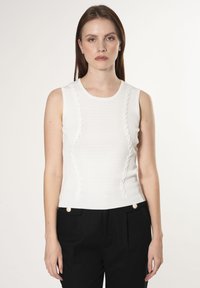 Top blanco sin mangas con acentos texturizados y ondulados en los lados, con un escote redondo y un diseño ajustado. Combinado con pantalones negros.