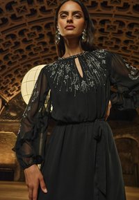 Čierne šaty s priesvitnými volánovými rukávmi, ozdobené precíznym strieborným kvetinovým vyšívaním a výstrihom v tvare kľúčovej dierky. Hladká, splývavá látka s opaskom v páse.