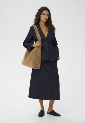 Naine, kes kannab tumedat teksastiilis bleiserit ja seelikut, hoides suurt helepruuni seemisnahast õlakotti, jalas mustad libisemiskingad, seisab ühevärvilise taustaga.