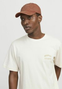 Brun cap med broderet "ORIGINALS STUDIO" og cremet kortærmet t-shirt. T-shirten har et lille gyldent logo på brystet.