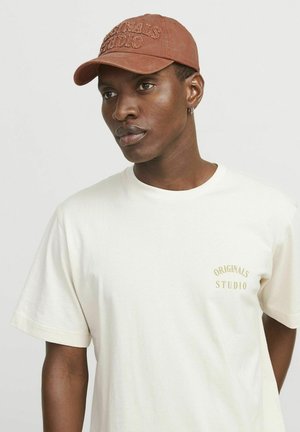 Brun cap med broderet "ORIGINALS STUDIO" og cremet kortærmet t-shirt. T-shirten har et lille gyldent logo på brystet.