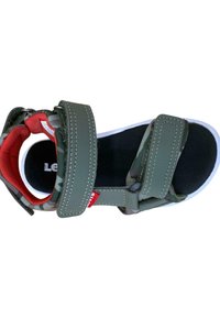 Sandalia verde con dos correas de velcro ajustables, forro acolchado rojo y planta negra texturizada. La suela blanca contrasta con el diseño superior.