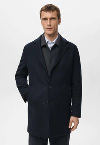 Abrigo de lana color azul marino con corte recto, diseño de un solo pecho, solapas con muesca y dos bolsillos delanteros. Se lleva sobre un suéter gris y una camisa azul.