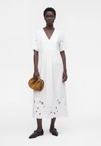 Femme portant une robe midi blanche avec des découpes florales, tenant une pochette tissée marron et portant des ballerines noires pointues, debout devant un fond blanc.