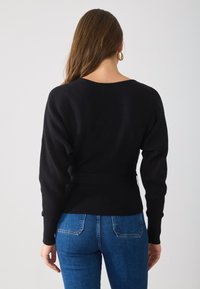 Schwarzer Strickpullover mit langen Ärmeln und weitem Ausschnitt, der eine figurbetonte Taille hat. Getragen über blauen Denim-Jeans mit Gesäßtaschen.