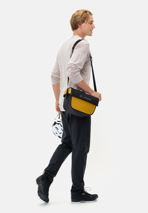 Borsa messenger nera e gialla con chiusura a patta, dotata di accessori in metallo e logo. Indossata a tracolla, con un casco bianco in mano.