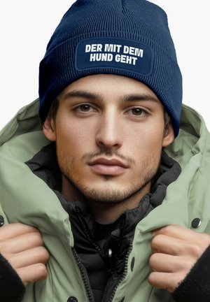 Navy ribbed knit beanie with a white patch that reads "DER MIT DEM HUND GEHT," worn with a green padded jacket.