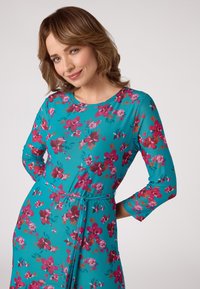 Bloemige jurk in turquoise, met 3/4 mouwen en een tailleband. Voorzien van een levendig patroon van roze en rode bloemen.