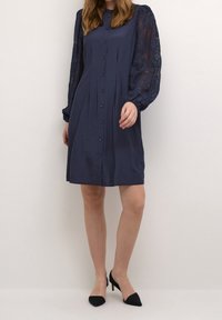 Mörkblå klänning med knappar framtill, långa spetsärmar och en avslappnad passform. Tyget har en slät yta som förstärker den eleganta designen.