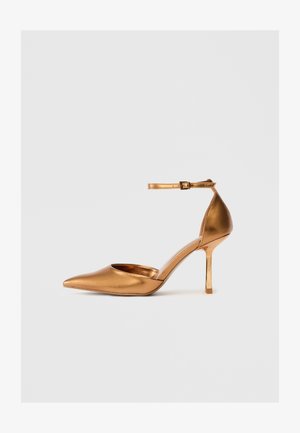 Sapato de salto alto metálico dourado com bico pontiagudo, lado aberto e tiracolo. Textura suave e acabamento brilhante. Salto stiletto de 5 polegadas.