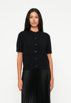 Kurzer schwarzer Cardigan aus weichem Material mit kurzen Ärmeln, rundem Ausschnitt und fünf schwarzen Knöpfen an der Vorderseite. Faltenrock in Schwarz darunter.