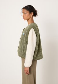 Žalias vilnonis liemenė su tekstūruota paviršiaus struktūra, plačiomis pažastimis ir mažu pleistru. Derinama su kreminiu megztiniu ir smėlio spalvos kelnėmis.