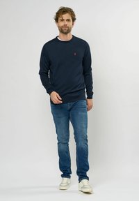 Dunkelblauer Strickpullover mit Rundhalsausschnitt, strukturiertem Stoff und kleinem roten Logo auf der Brust; kombiniert mit hellblauen Jeans und weißen Sneakers.
