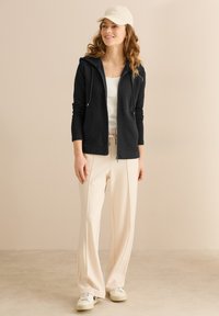 Sudadera negra con cremallera y cordones, top beige sin mangas, pantalones de pierna ancha color crema y zapatillas deportivas blancas, todo sobre un fondo neutro.