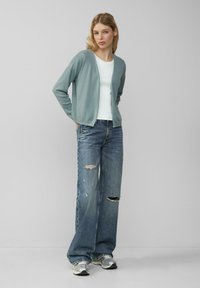 Hellblauer Cardigan aus weichem Stoff, kombiniert mit einem weißen Oberteil und lässigen, weit geschnittenen Jeans. Silberne Sportschuhe runden den Look ab.