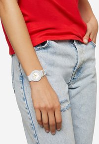 Hvidt ur med en rund urskive og guldfarvede detaljer, udstyret med en grå silikonesnor og en stjerneformet charms. Båret på en hånd ved siden af lyseblå jeans.