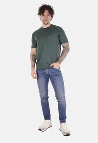 T-shirt verde a maniche corte, collo rotondo, indossato con jeans blu slim-fit e sneaker chiare. Tessuto in cotone con una texture liscia.