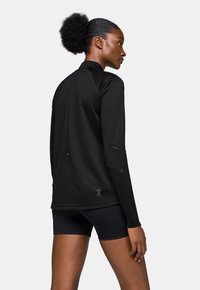 Haut de sport noir à manches longues avec col montant, tissu texturé et détails réfléchissants, associé à un short noir.