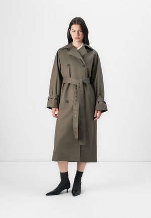 Oliivinvihreä trench-coat, jossa on kaksirivinen etuosa, vyötärölle tuntuvat vyöt ja niittitiedot. Pitkät hihat ja midi-pituus, yhdistetty mustiin kenkiin.