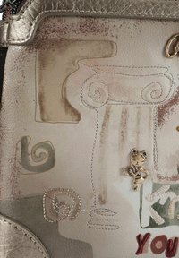 Pelle cremosa strutturata con motivi astratti, caratterizzata da un profilo decorativo a colonna e un ciondolo di metallo a forma di gatto. Dettagli cuciti in marrone e argento.