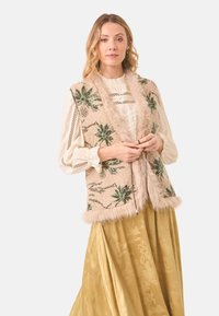 Chaleco de piel sintética beige con bordado floral verde, rematado con suave piel en los bordes, usado sobre una blusa crema y una falda dorada.