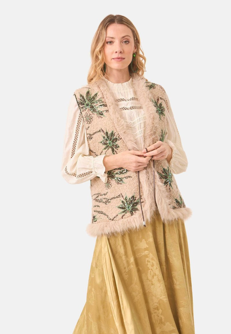 Chaleco de piel sintética beige con bordado floral verde, rematado con suave piel en los bordes, usado sobre una blusa crema y una falda dorada.