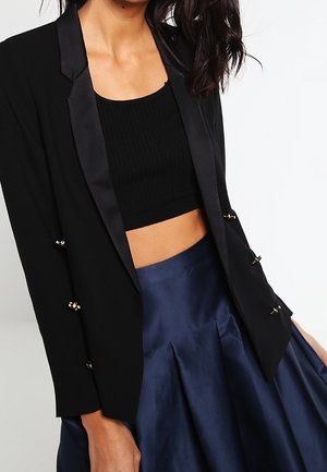 Vrouw die een zwart cropped tanktop draagt, een zwarte blazer met satijnen revers en een hoog opgesloten marineblauwe geplooide rok.