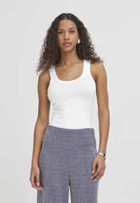 Jonge vrouw met krullend haar, gekleed in een wit tanktop, grijze broek met patroon, zilveren creolen en een zilveren armband, staand tegen een effen achtergrond.