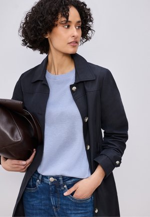 Mujer con cabello rizado que lleva un abrigo negro sobre un suéter azul claro y jeans, sosteniendo un bolso grande de cuero marrón oscuro bajo el brazo.