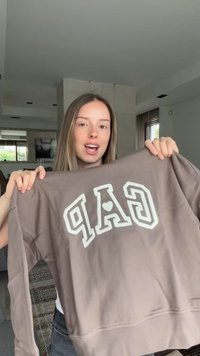 Jeune femme tenant un sweat marron avec des lettres blanches "GAP" et un symbole cœur au centre, à l'intérieur dans un salon.