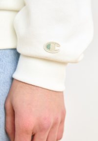 Krémszínű pulóver anyag mutatva egy csuklón, kis hímzett logóval zöld és bézs részletekkel. Simaság jól látható.