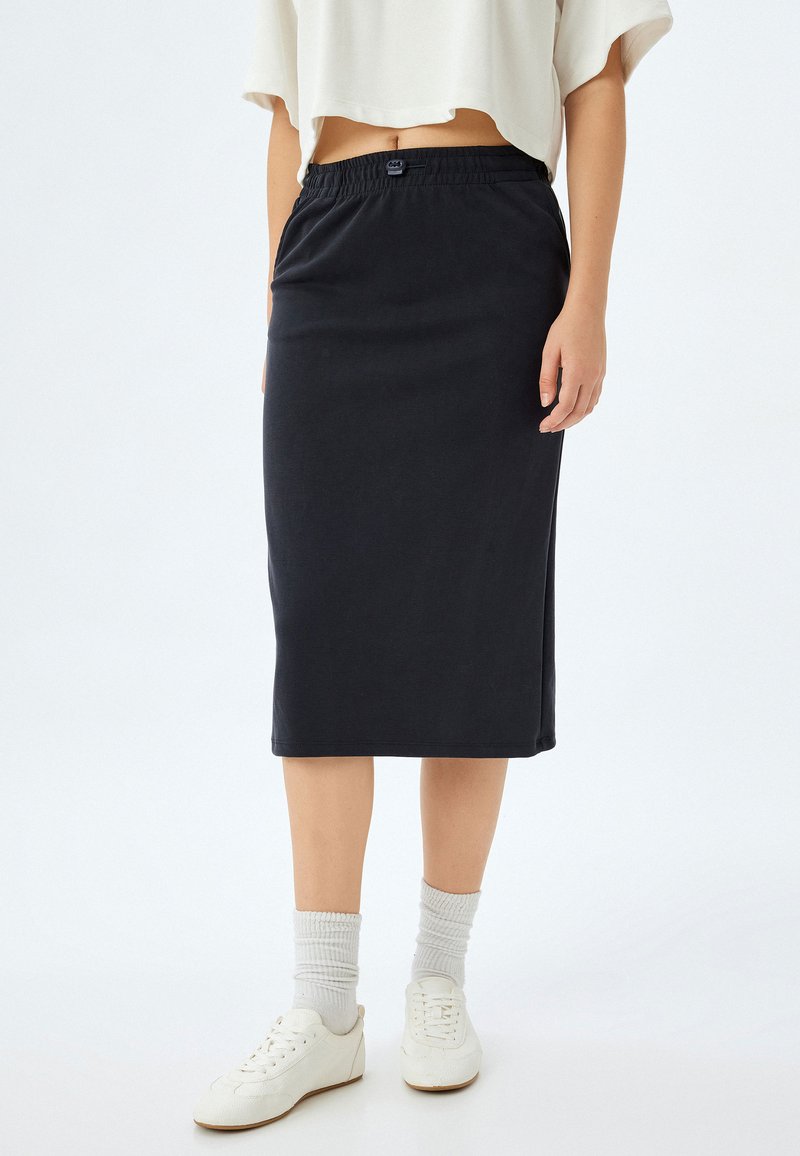 Zwarte katoenen midi-jurk met een elastische tailleband, met een gladde textuur en een ontspannen pasvorm, gecombineerd met witte schoenen en grijze sokken.