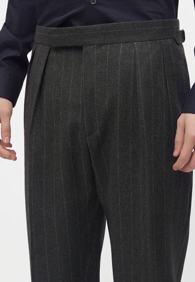 Pantalon gris foncé à fines rayures avec plis, porté avec une chemise bleu marine foncé rentrée, montrant les mains sur les côtés et une taille cintrée.
