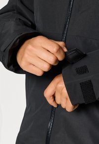 Melnā ūdensizturīgā jaka ar elegantu dizainu. Aprīkota ar regulējamiem manžetēm ar Velcro aizdari un redzamu priekšējo rāvējslēdzēju. Tekstūras auduma detaļa.