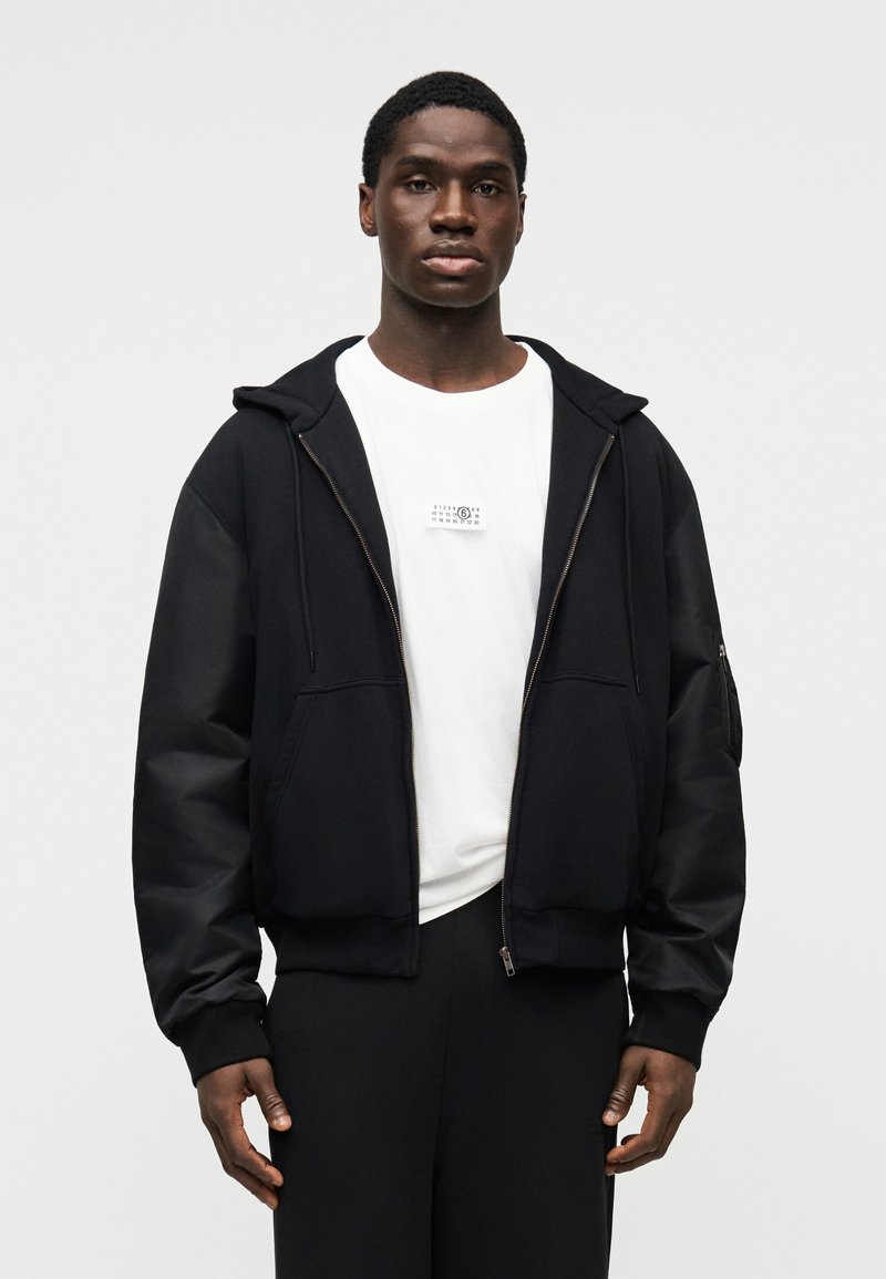 Sort hættejakke med lynlås og store lommer foran samt hætte. Lagdelt over en hvid t-shirt med et lille trykt motiv.