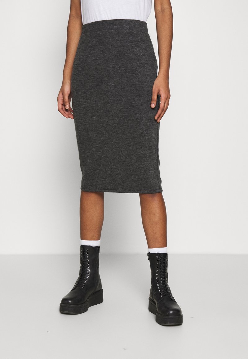 Femme portant une jupe crayon gris foncé longueur genou, des chaussettes blanches et des bottines noires à lacets, debout devant un fond clair uni.