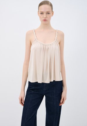 Lichtbeige mouwloze top met gathered stof, elastische halslijn en gevlochten bandjes, gecombineerd met donkere denim wijde broekspijpen.