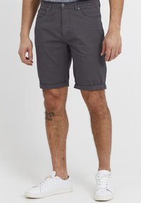 Mann i mørkegrå rullede shorts, hvite sneakers og synlig tatovering på venstre bein, som står mot en ensfarget bakgrunn.