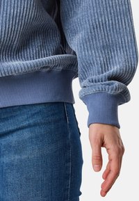 Felpa blu a costine con polsini e orlo elasticizzati, abbinata a jeans in denim blu scuro. Focalizzati sulla texture e sui dettagli dei tessuti sovrapposti.