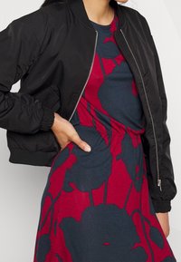 Veste bomber noire avec fermeture éclair, poignets côtelés et ourlet élastique, portée sur une robe bleue ornée de grands motifs floraux rouges.