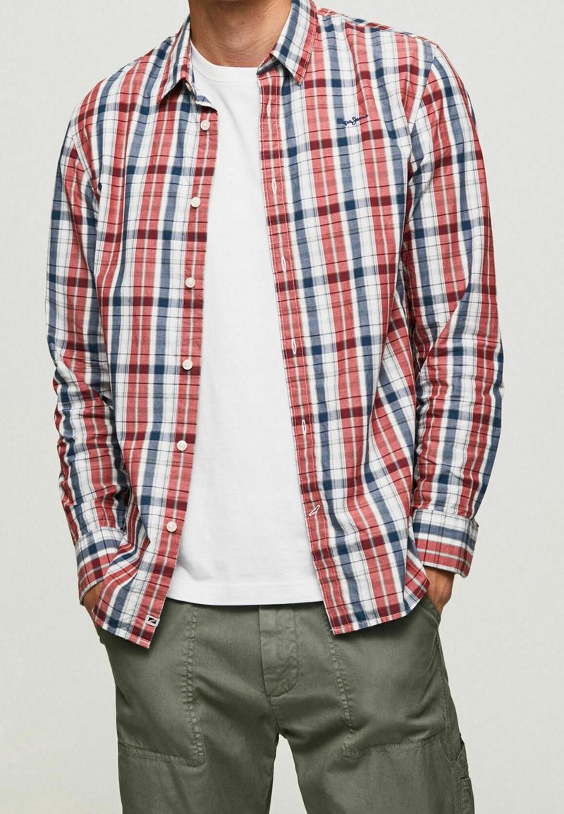 Homme portant une chemise à carreaux rouge, blanche et bleue boutonnée sur un t-shirt blanc avec un pantalon cargo vert, mains dans les poches.