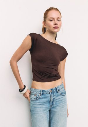 Mlada žena s crvenom kosom, odjevena u usku smeđu kratku majicu i svijetloplave traperice, pozira s jednom rukom u džepu na jednostavnoj pozadini.