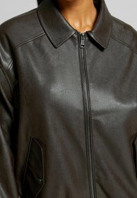 Brązowa kurtka z sztucznej skóry z kołnierzem, zamek błyskawiczny z przodu oraz dwiema bocznymi kieszeniami ozdobionymi guzikami. Gładka tekstura.