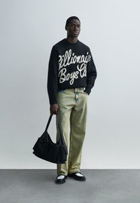 Μαύρο πλεκτό πουλόβερ με λευκό κείμενο, ανοιχτοπράσινο παντελόνι κοτλέ και μαύρη τσάντα. Τα παπούτσια είναι μαύρα και λευκά με στρογγυλεμένη μύτη.