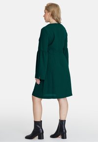 Rochie verde, până la genunchi, cu mâneci clopot și talie ajustată, asortată cu botine negre. Design simplu, fără modele vizibile.