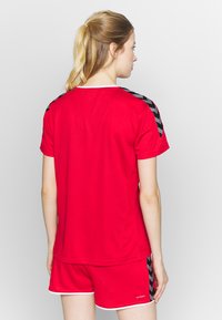 Rød atletisk t-shirt med korte ærmer, der har sorte og hvide chevron-accenter på skuldrene. Kombineret med røde shorts og hvid kant.
