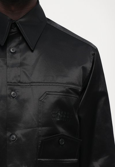 Mustmust shirt, mis glansmaterjalist, millel on terav krae, eesmine tasku nuppude sulgemisega ja peened õmbluste detailid.
