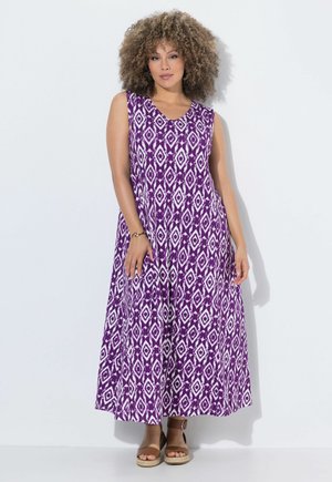 Mujer con cabello rizado que lleva un vestido maxi sin mangas con estampado púrpura y blanco y sandalias de cuña marrones, de pie contra una pared blanca.