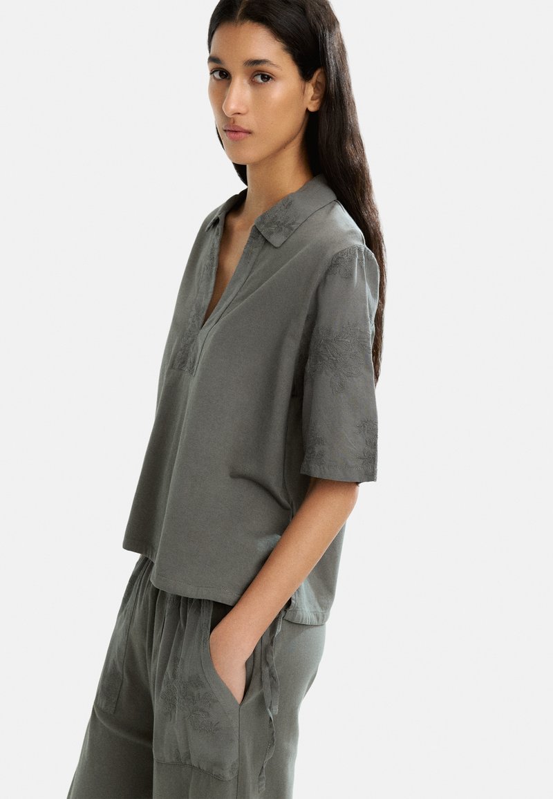 Blusa de manga corta, gris, con cuello de solapa y un sutil patrón floral en las mangas, hecha de una tela suave y con un corte relajado.